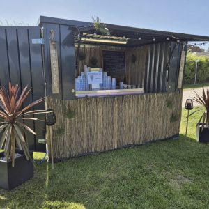 Mobiler Verkaufscontainer mit Bambusverkleidung und Theke, dekoriert mit Pflanzen und Palmen. Beschilderung ‚Heimathafen‘ und Getränkekarte für Cocktails.